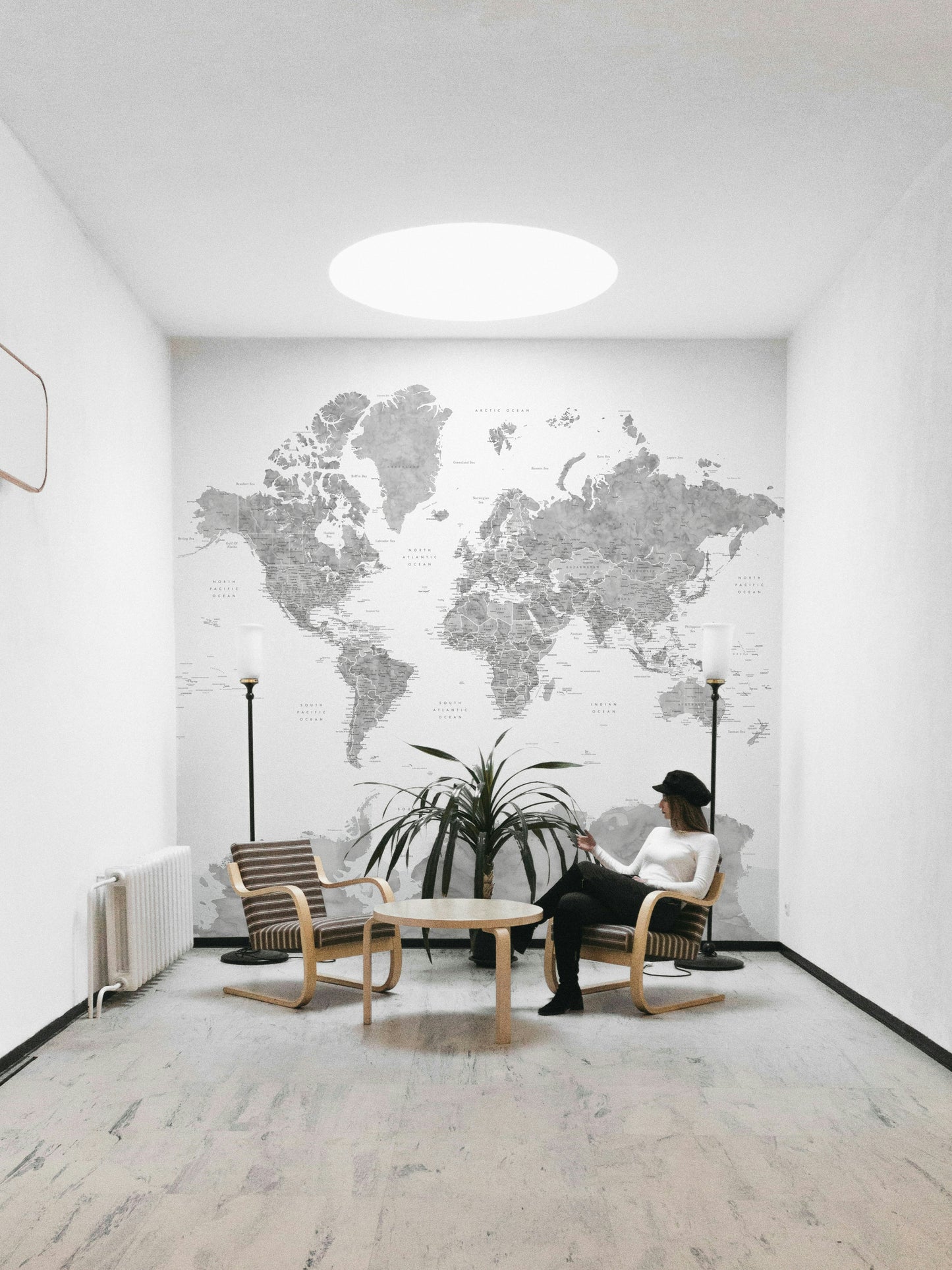 Gray Watercolor World Map wallpaper mural in soft gray behind a woman seated in a wooden armchair