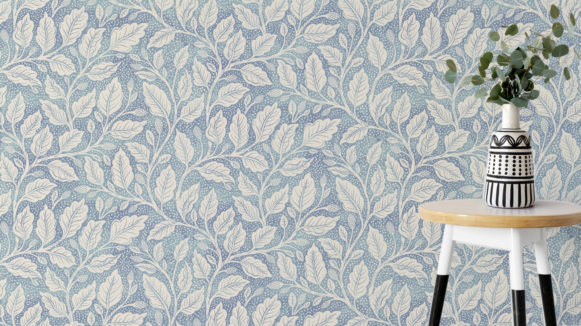 Blue Budding Branches wallpaper with cream leafy vines on dusty blue behind a white round stool and patterned vase