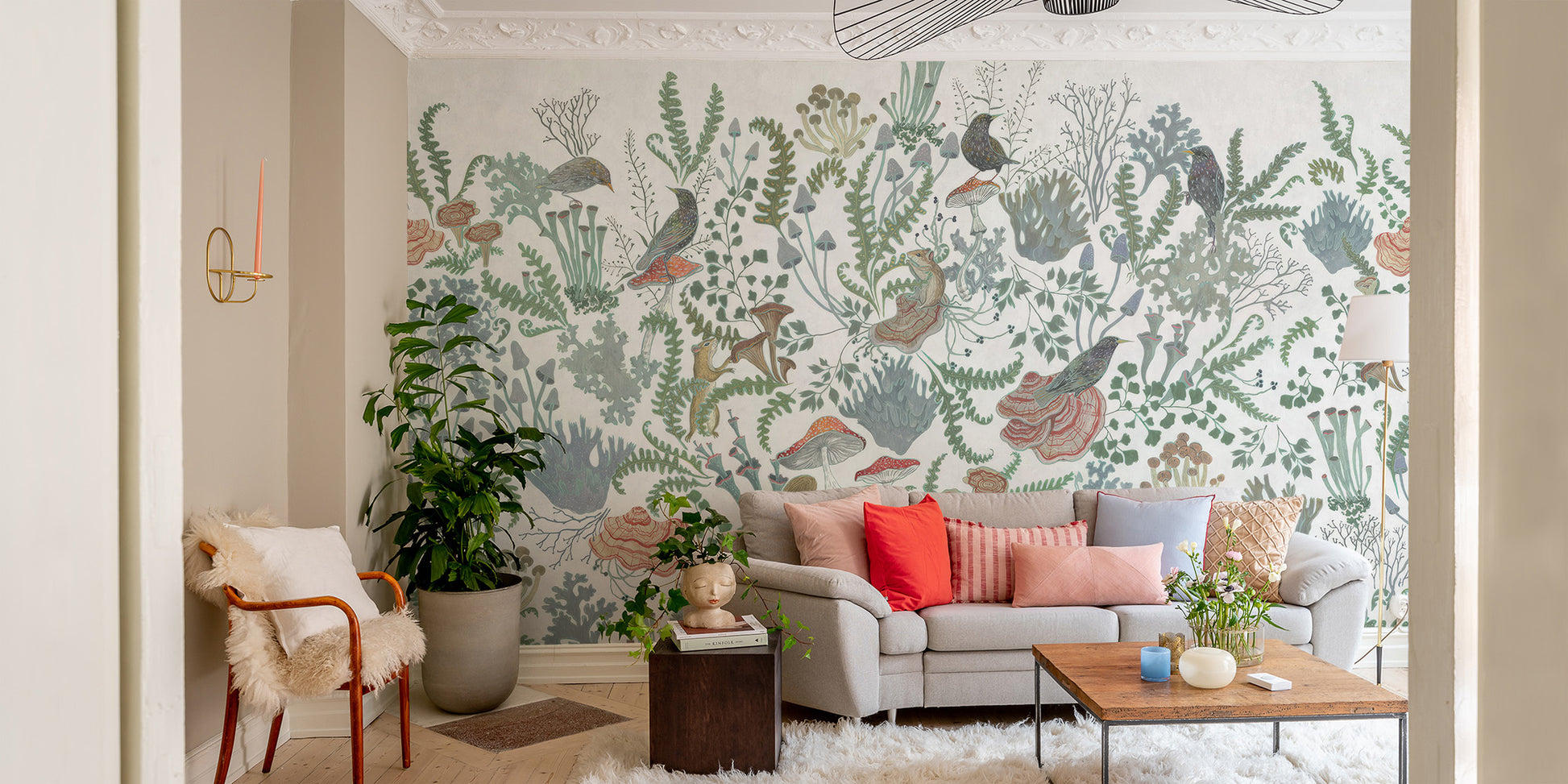 Botanical Nest Fresco wallpaper mural of green foliage and birds behind a gray sofa with coral pillows in a living room