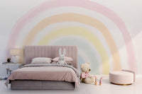 Pastel Rainbow wallpaper mural behind a blush pink upholstered bed with bunny plush and teddy bear in a soft bedroom