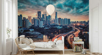 Cityscape Dawn wallpaper mural of the dusk city skyline above a white bed and rattan chair in a bedroom