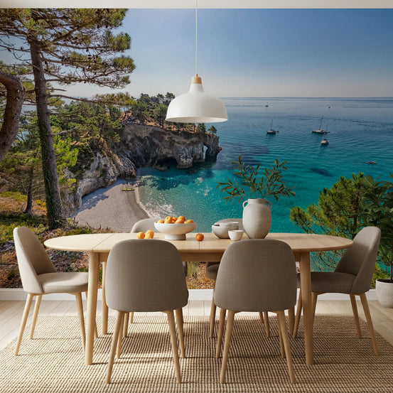 Coastal Haven wallpaper mural of a turquoise cove with pines and sailboats behind an oval wooden dining table and taupe chairs