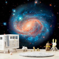 Cosmic Symphony wallpaper mural with a blue and orange spiral galaxy above a white crib in a nursery with plush toys