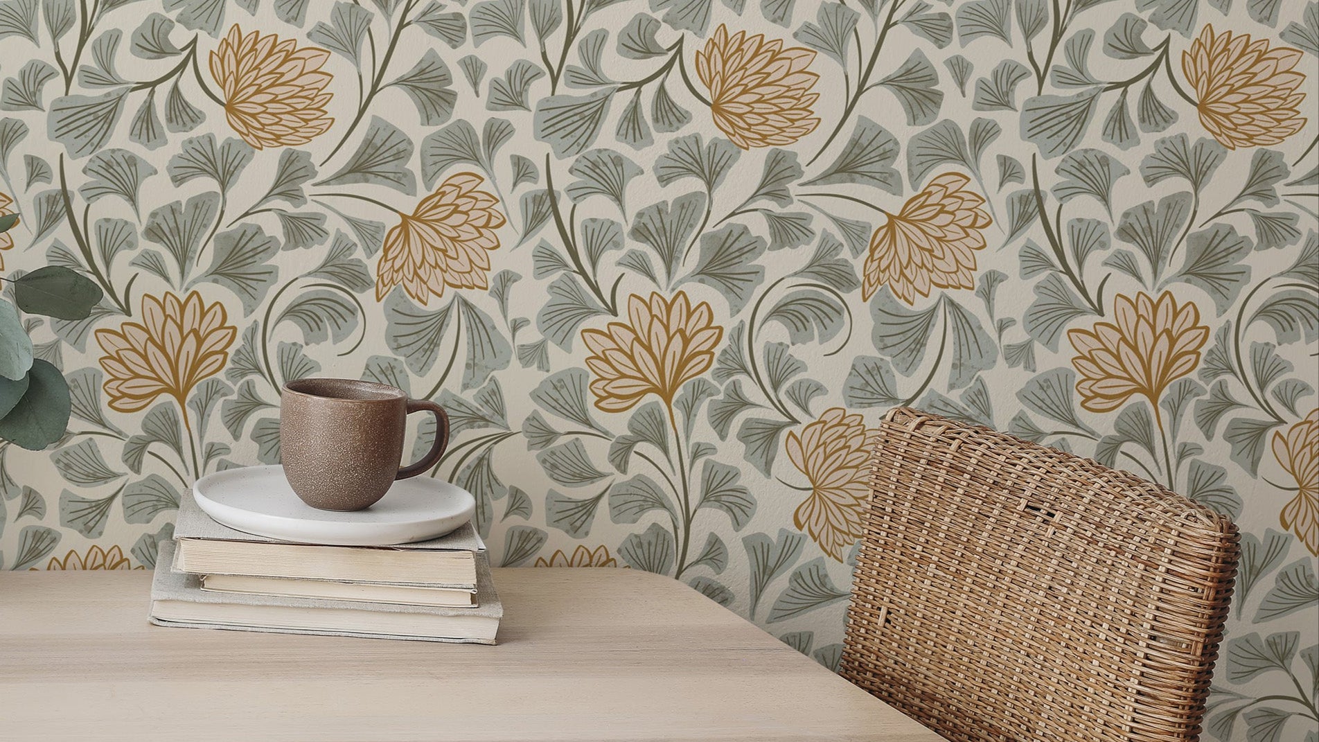 Dancing Bloom wallpaper with mustard florals and sage leaves behind a stacked books and brown mug on a wood desk