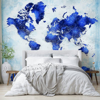 Deep Blue Watercolor World Map wallpaper behind an upholstered bed with white duvet and grey knit throw