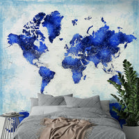 Deep Blue Watercolor World Map wallpaper behind a grey tufted bed with a beige throw and blue side table