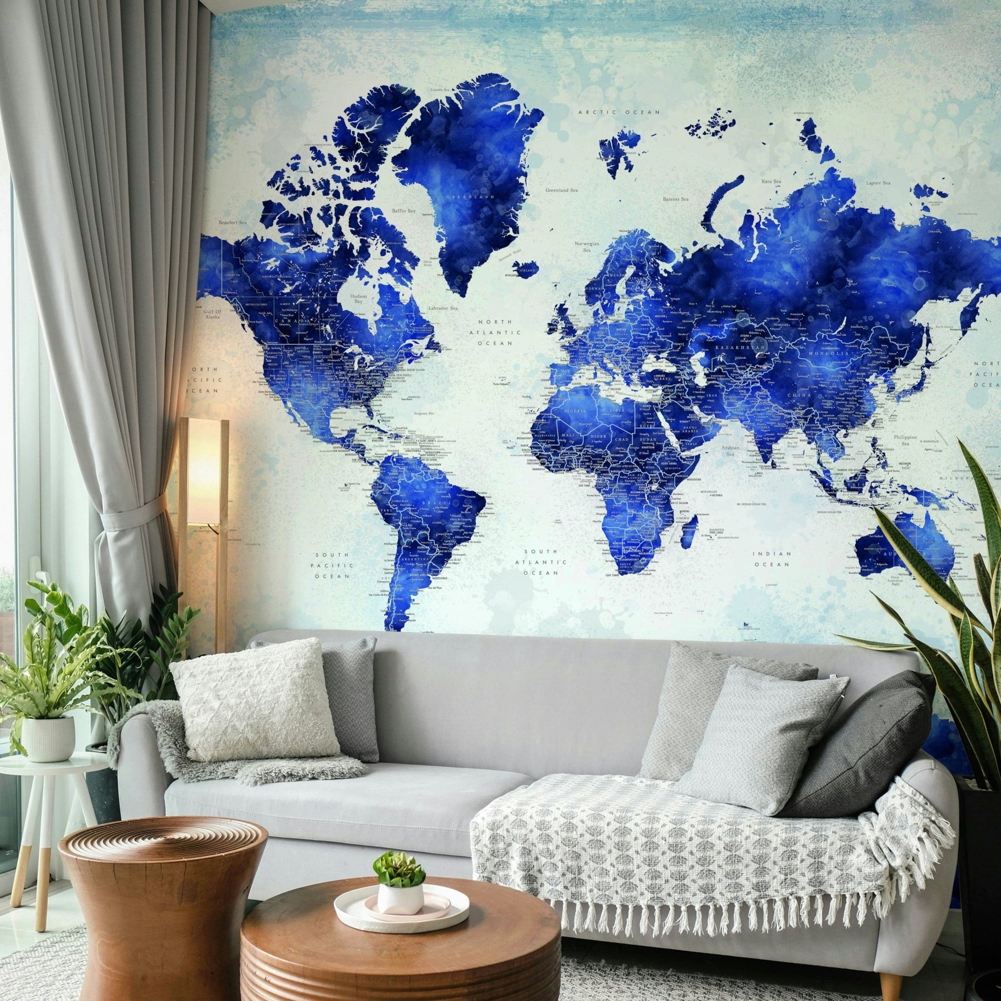 Deep Blue Watercolor World Map wallpaper behind a light grey sofa with a floor lamp and wood drum tables
