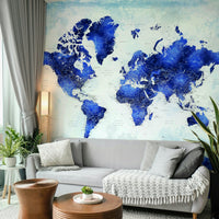 Deep Blue Watercolor World Map wallpaper behind a light grey sofa with a floor lamp and wood drum tables