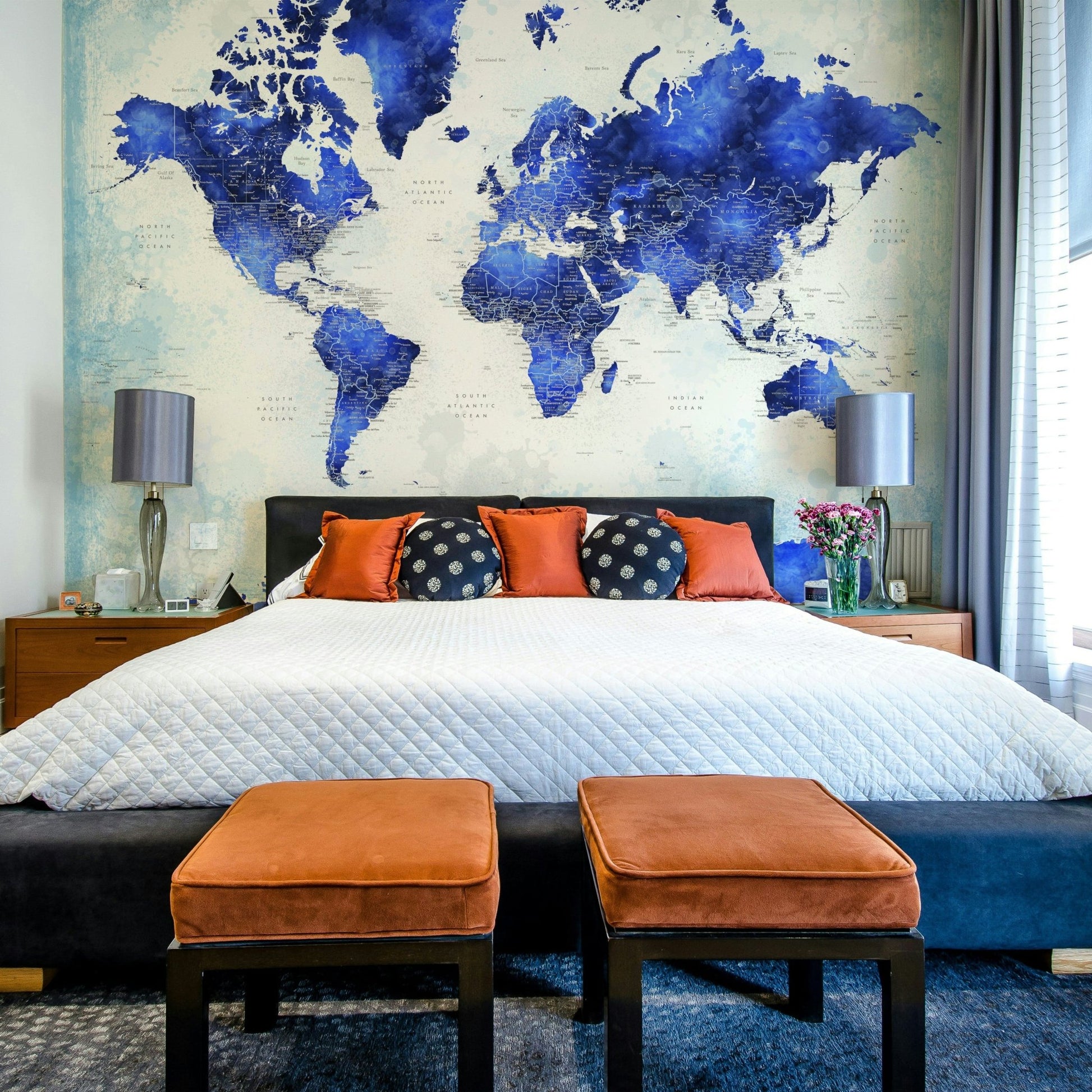 Deep Blue Watercolor World Map wallpaper as a headboard above a bed with orange pillows and ottomans