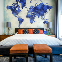 Deep Blue Watercolor World Map wallpaper as a headboard above a bed with orange pillows and ottomans