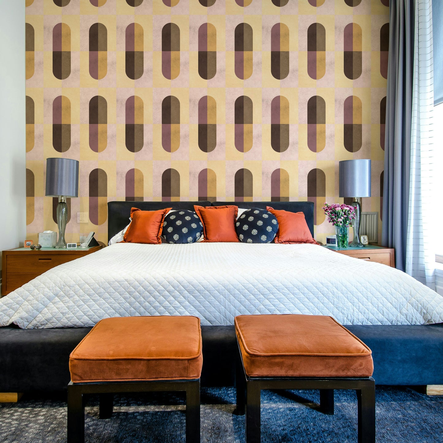 Earthy Geometric wallpaper behind a bed with a black headboard, orange cushions and twin grey lamps