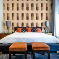 Earthy Geometric wallpaper behind a bed with a black headboard, orange cushions and twin grey lamps