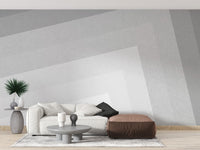 Geometric gray ombre layered wallpaper mural with angled tones behind a white sofa and leather ottoman in a living room