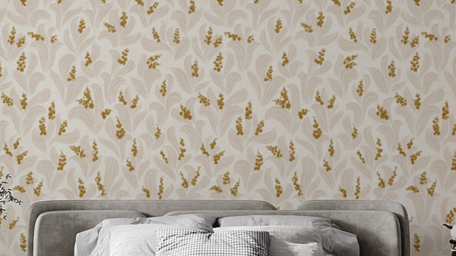Golden Whispers Lily Valley wallpaper in cream with golden lily sprigs above a grey upholstered headboard in a bedroom