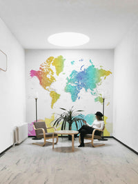 Rainbow World Map wallpaper in bright watercolor behind a woman in a black hat seated between two floor lamps in a room