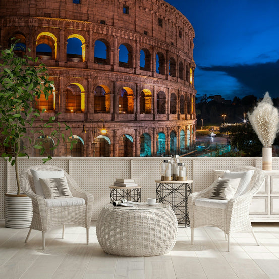 Roman Nights wallpaper mural of the lit Colosseum at night behind white wicker chairs on a patio terrace
