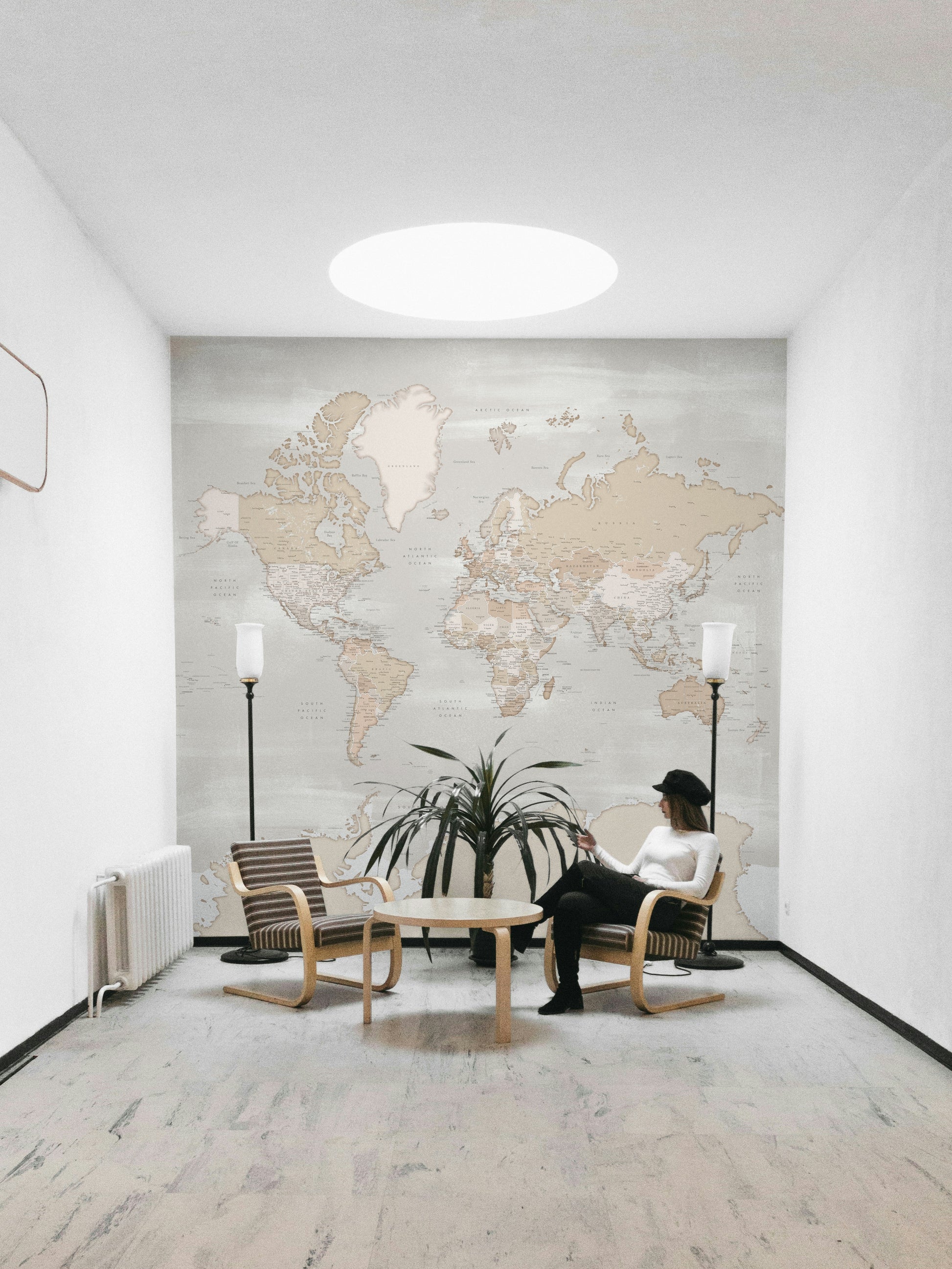 Rustic World Map wallpaper in beige behind a woman in a black hat seated between two floor lamps in a bright room
