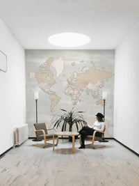 Rustic World Map wallpaper in beige behind a woman in a black hat seated between two floor lamps in a bright room