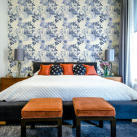Serene Blue Botanicals wallpaper above a black bed with white quilt and orange velvet stools in a bedroom