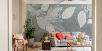 Elegant Teal Botanical Leaf wallpaper mural with white leaves behind a grey sofa with coral pillows in a living room