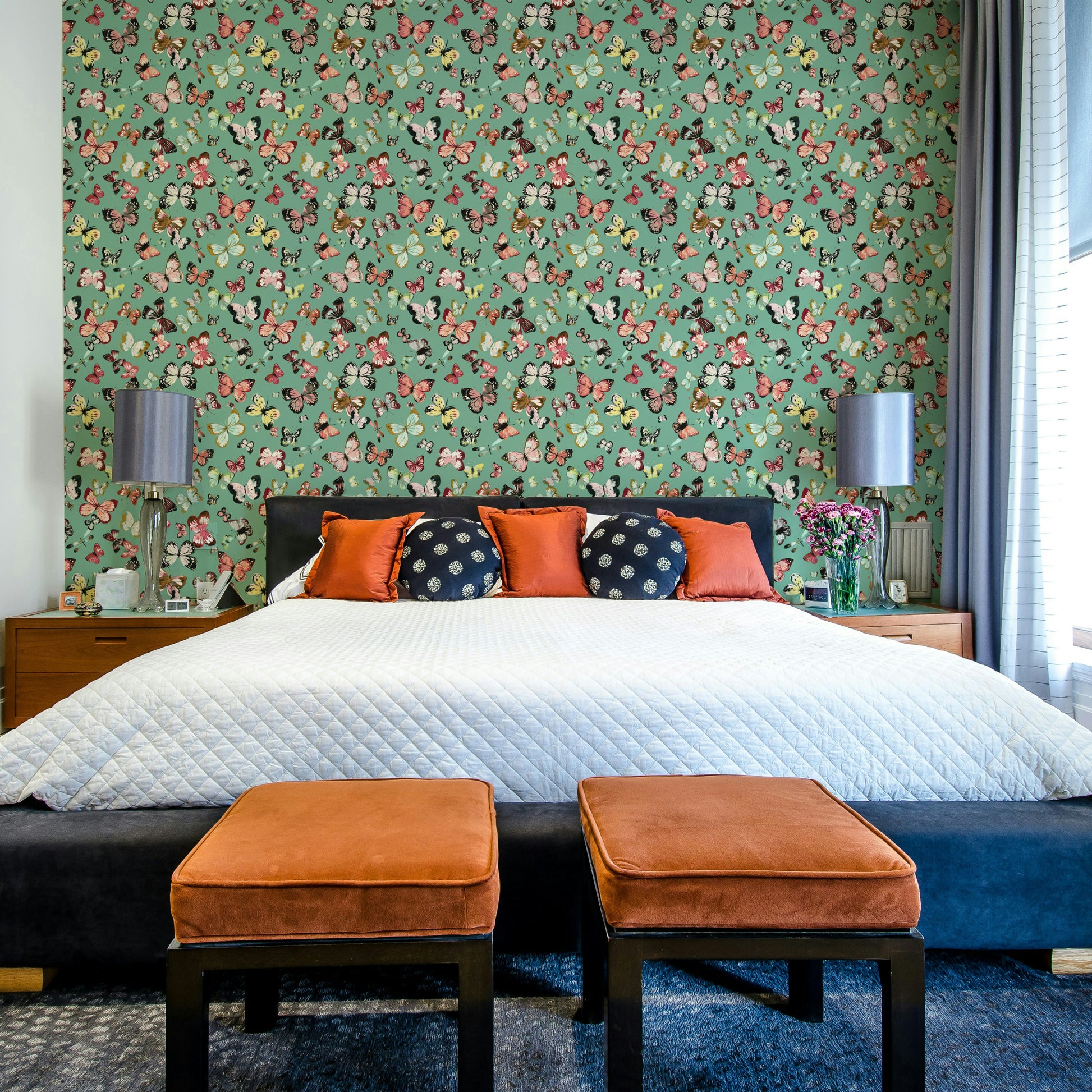 Flying Butterflies Colorful Teal Coral wallpaper above a bed with orange and navy pillows and velvet ottomans