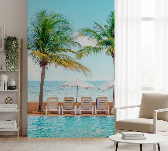 Turquoise Escape wallpaper mural of a palm-lined resort pool behind a beige armchair and white shelving in a living room