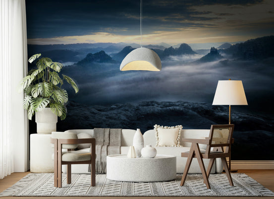 Twilight Mountain Dream wallpaper mural of misty blue peaks above fog behind a cream sofa, wood chairs and a monstera in a living room