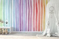 Watercolor Rainbow Striped wallpaper mural with bright rainbow stripes behind a green armchair and white rocking horse in a kids room